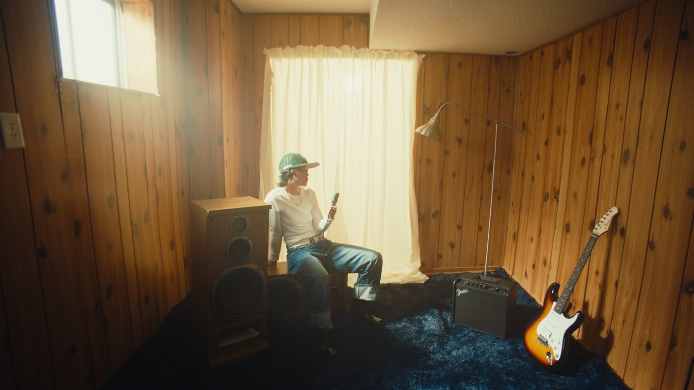 A musician sits in a wood-panelled basement, backlit by a curtained window
